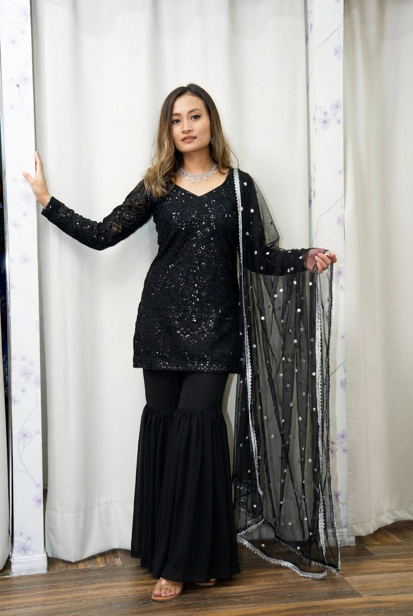 Woman wearing a black traditional outfit with a sheer dupatta against a white curtain background