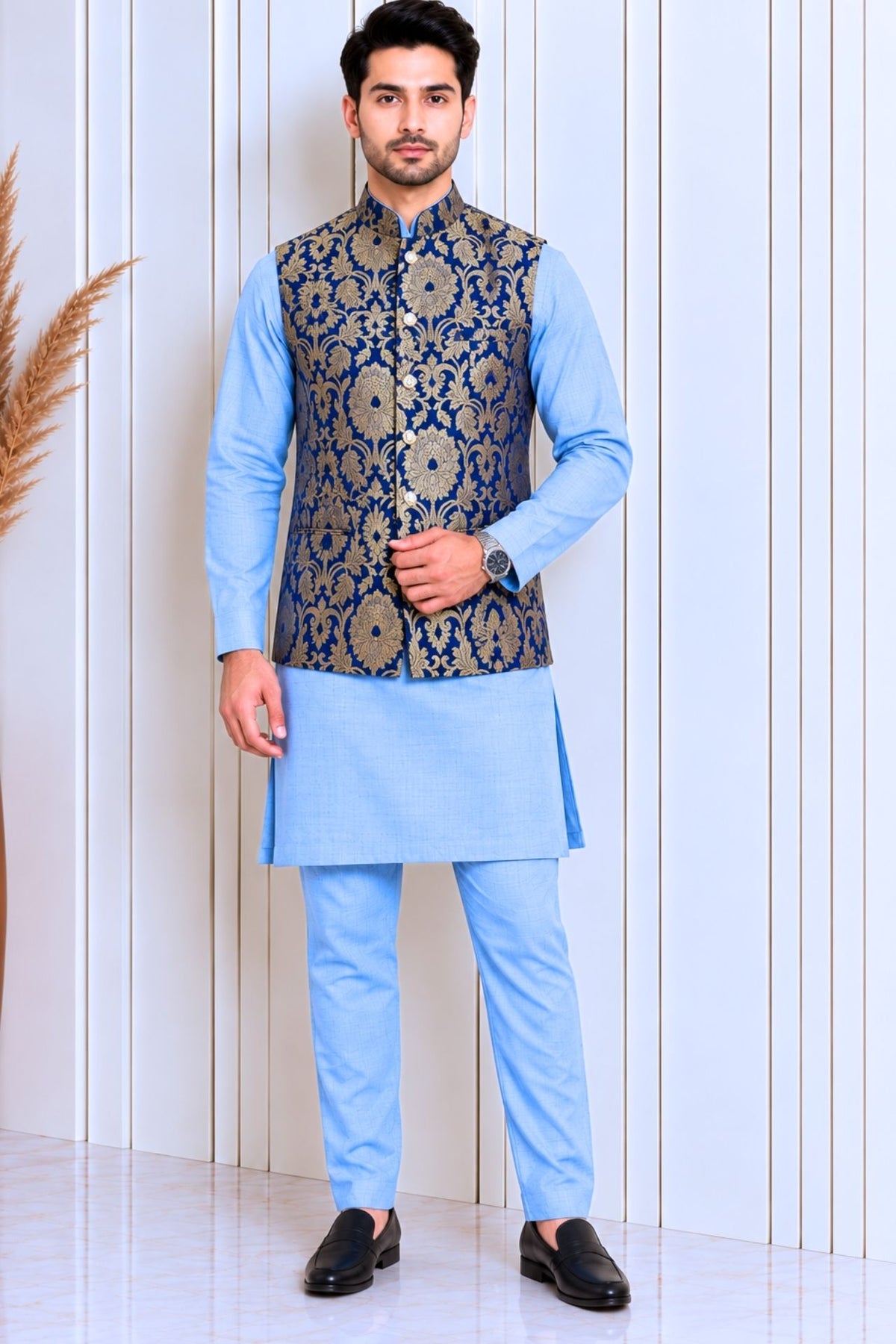 A man modeling a light blue traditional Indian outfit consisting of a kurta, trousers, and a navy blue waistcoat with gold floral embroidery, standing against a white paneled wall.